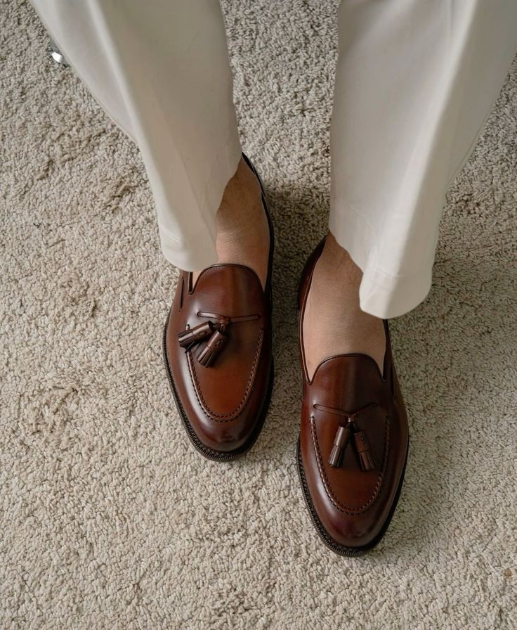 Brown loafers with tassels worn with white pants on a beige carpet.