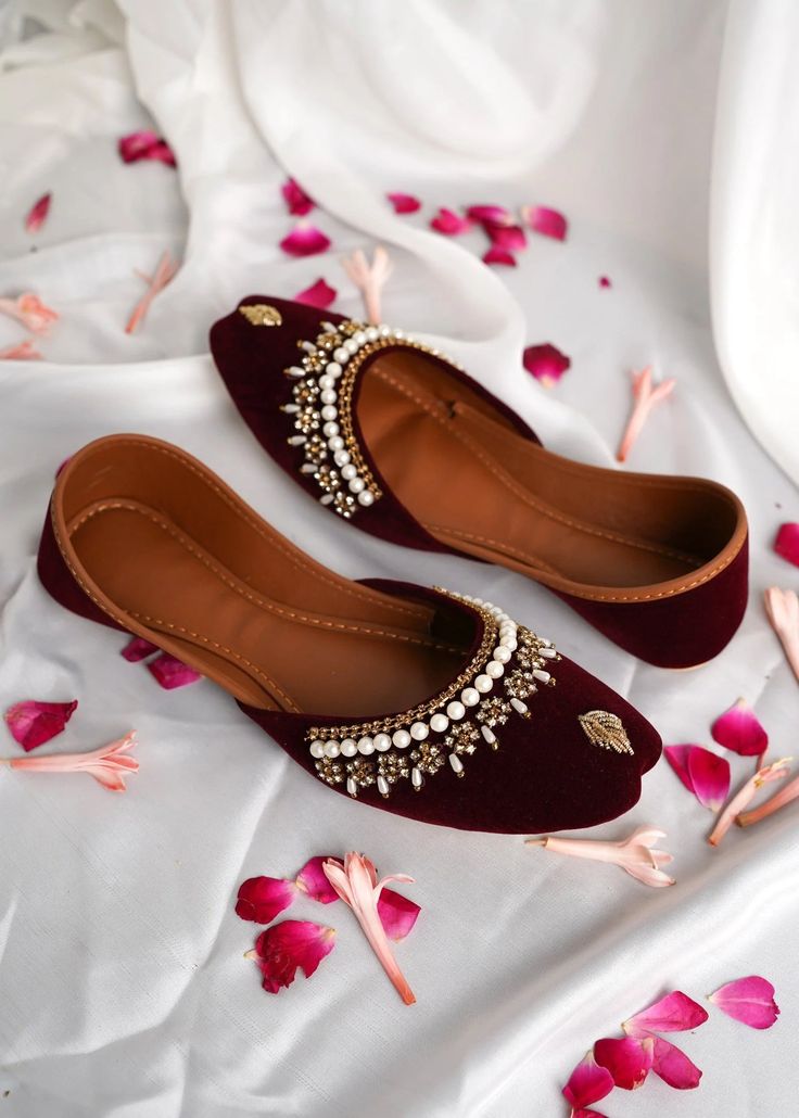 Pair of maroon flats with gold embellishments on a white fabric background with pink petals.