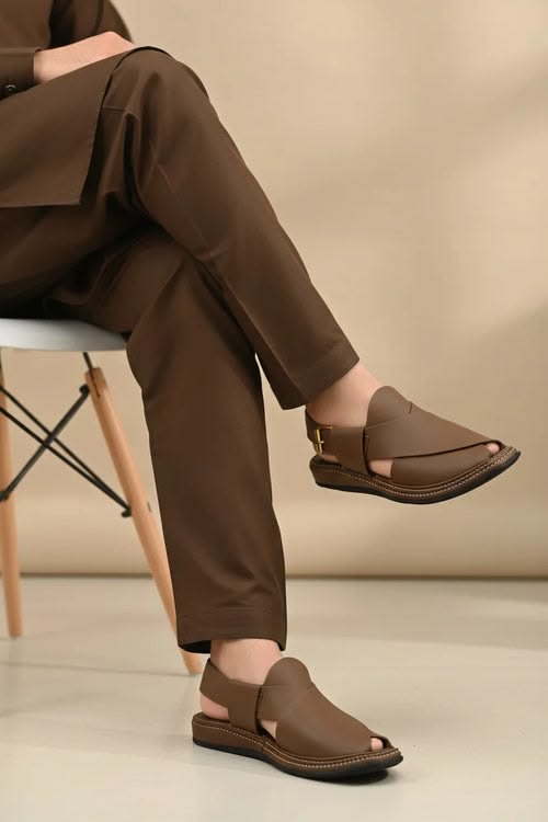 Person wearing brown sandals and pants sitting on a chair with a neutral background