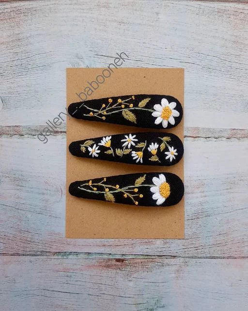 Three black hair clips with floral designs on a beige card, placed on a textured white surface.