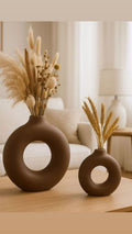 Two brown circular vases with dried plants on a wooden table in a living room setting.