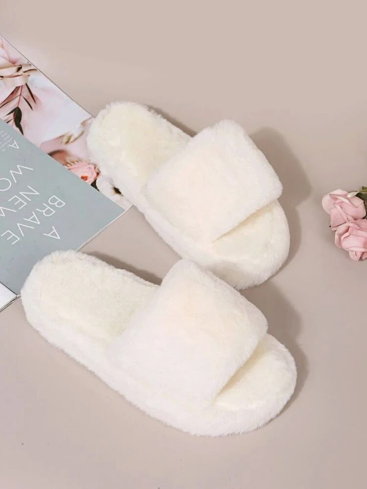 White fluffy slippers on a beige surface with a magazine and flowers in the background.